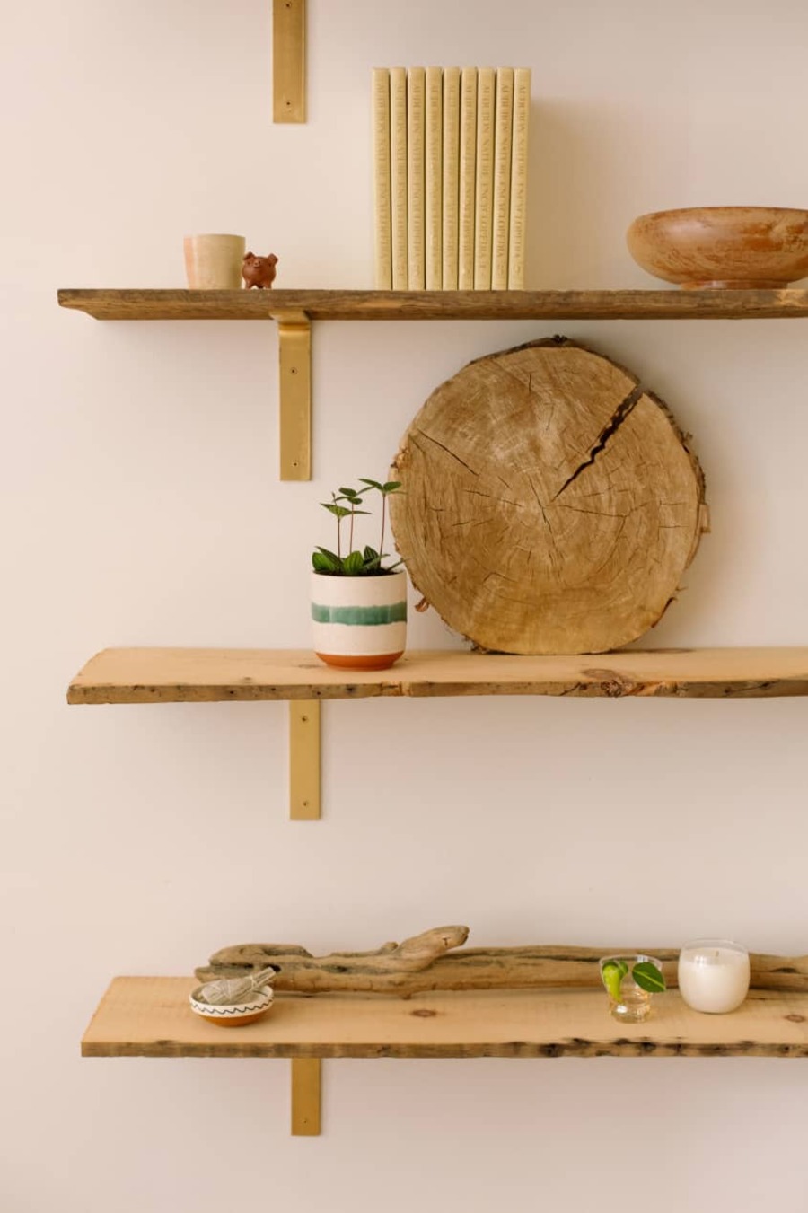 Repisa de madera rústica de tres niveles con diferentes elementos de madera y pequeños adornos en cada uno, incluyendo plantas, una vela, un bowl, libros y una parte de un tronco