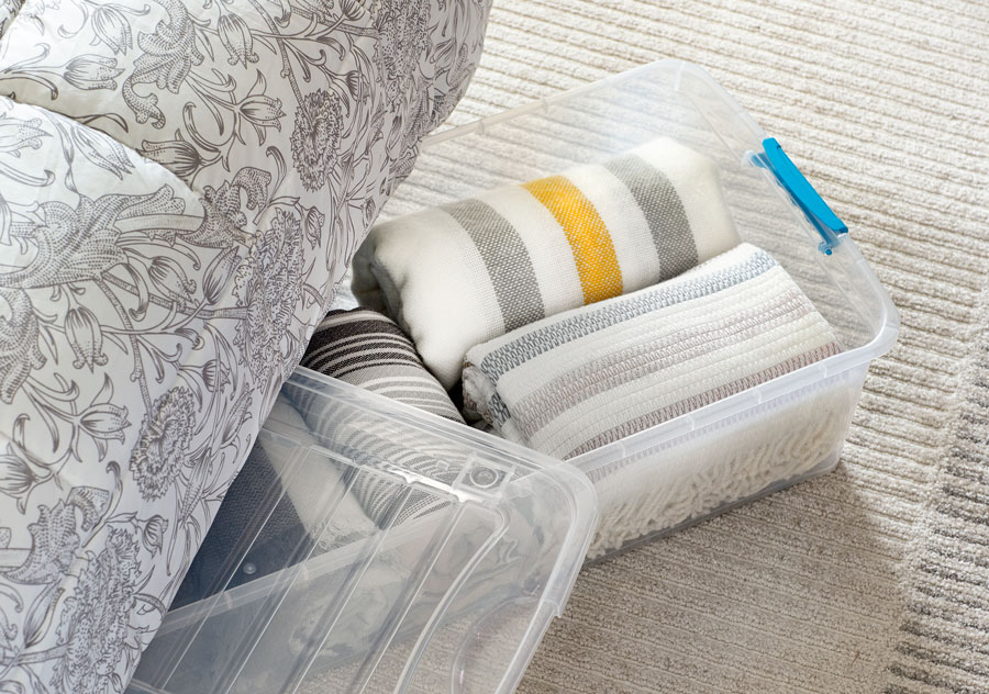 Caja plástica con mantas en tonos neutros asomándose debajo de una cama.